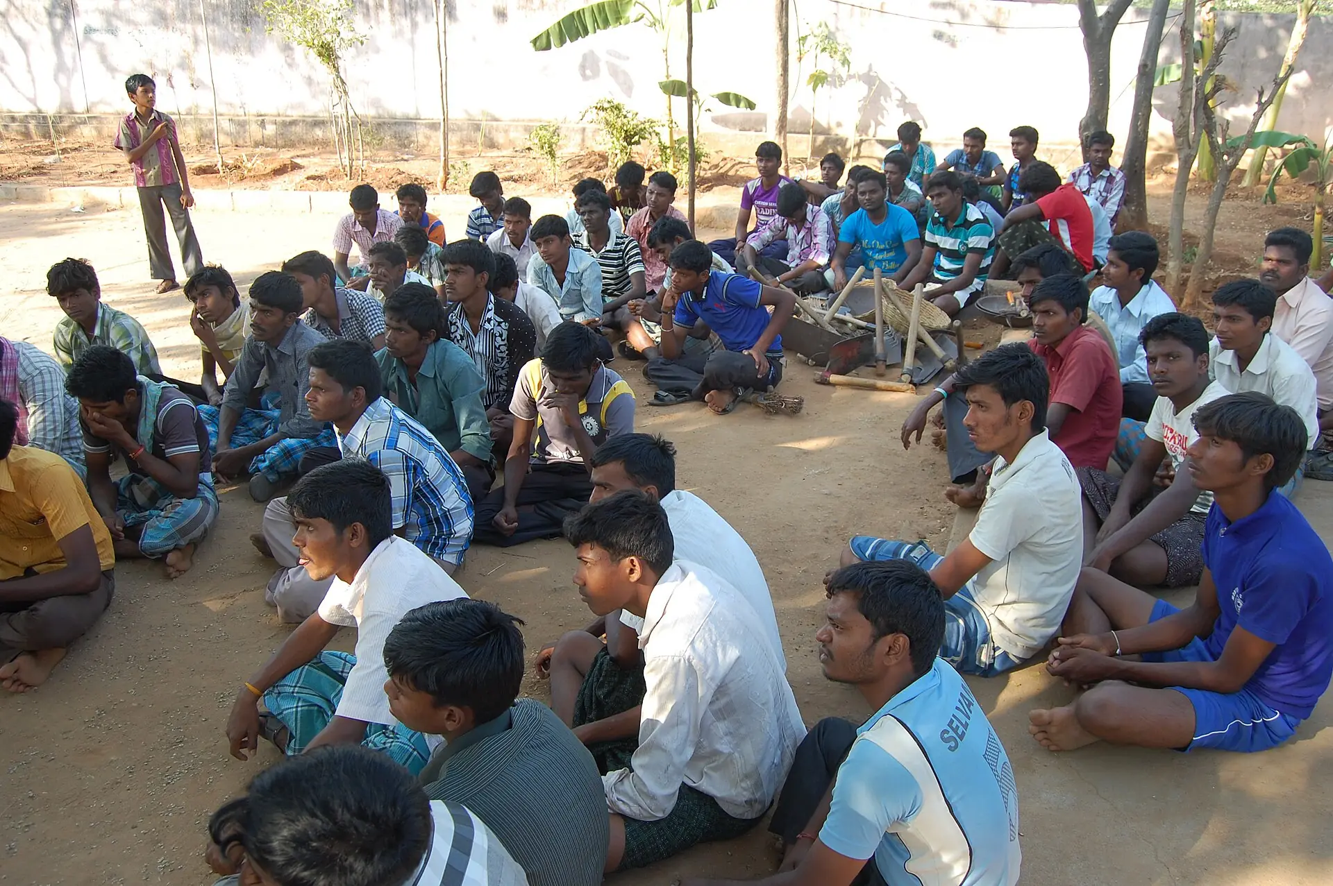 A large group of young men seated on the ground under trees, listening to a training session